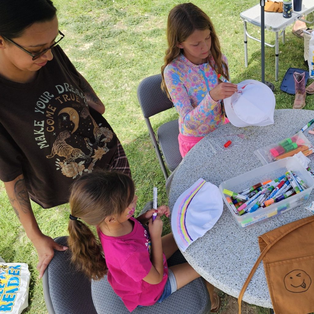two young girls decorate hats with fabric pens at a craft table.