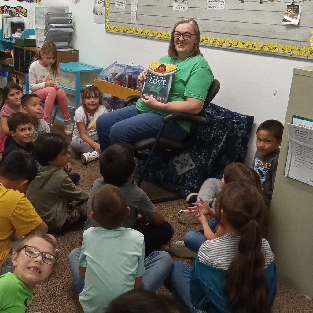 A teacher holds up the children's book she will be reading to the class of students sitting cross-legged at her feet.