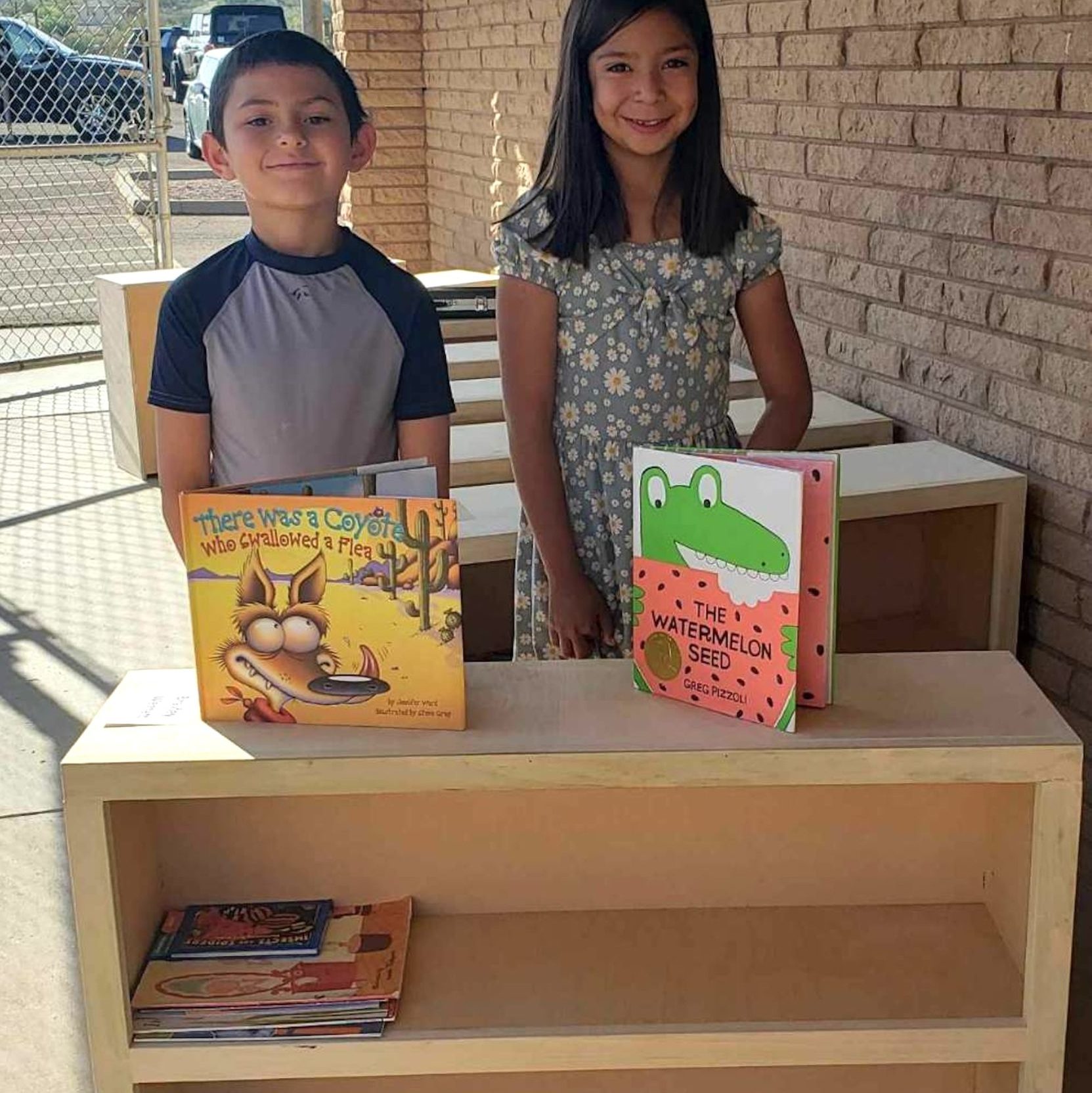 Two students smile proudly with children's books displayed on top of a beautiful wood bookshelf.